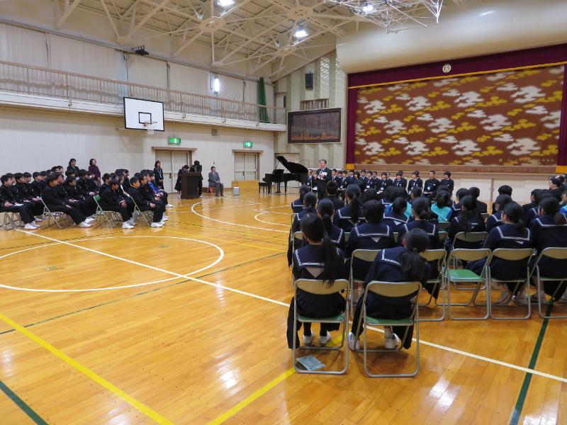 体育館で縦2列横並びでパイプ椅子に座る陸前高田市の中学生たちの前で右手にマイクを持った男性教師がその前方両端に斜めに縦4列で横並びにパイプ椅子に座る生徒たちの方を向きながら喋っている写真