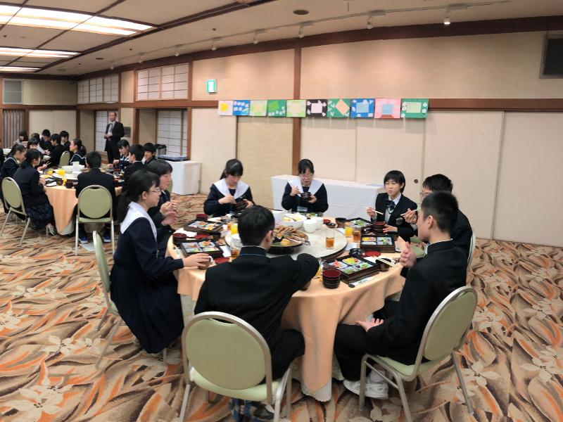 数人一グループで円卓テーブル席に座りながら各々出された弁当を食べながら会話をする中学生たちとその様子を前方で見守る男性の写真
